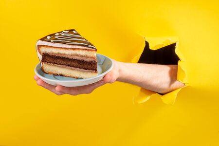 Hand holding a big piece of chocolate cake through a torn hole in yellow paper