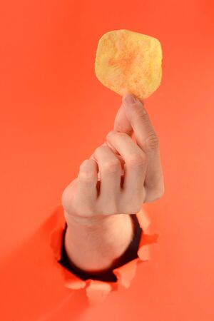 Hand holding a potato chip through a torn hole in red paper background.の写真素材