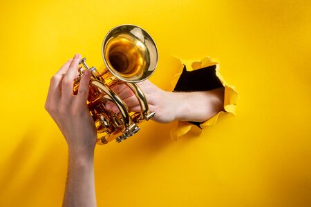 Hands playing pocket trumpet through a torn hole in yellow paper background. Jazz music event advertisement concept.の写真素材