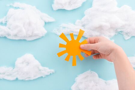 Child holding a cut-out orange paper sun on blue sky with cotton wool cloudsの写真素材