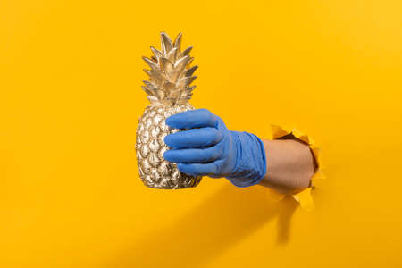 Hand in a latex glove holding a golden pineapple through torn yellow wall