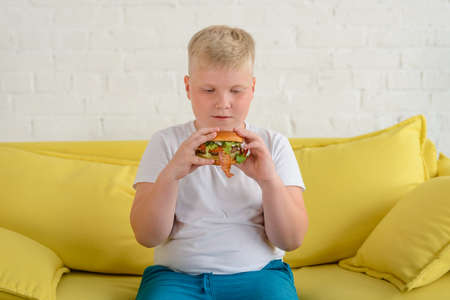 Obese boy eating a hamburger, sitting on a yellow couch