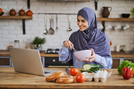 Muslim woman mixing ingredients in bowl and watching something funny on a laptopの写真素材
