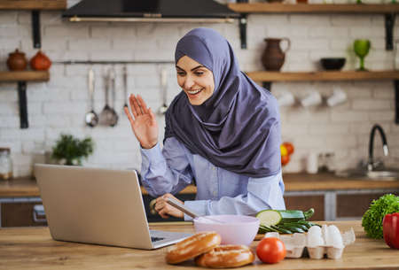 Arabian woman waving during a video call on a laptop and cooking in the kitchenの写真素材