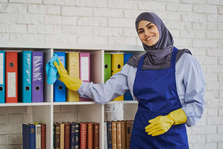 Cheerful Arabian woman wiping dust off the bookshelvesの写真素材