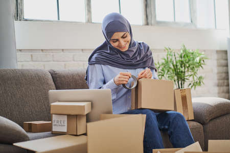 Joyful Muslim woman packaging items into boxes and putting adhesive tapeの写真素材