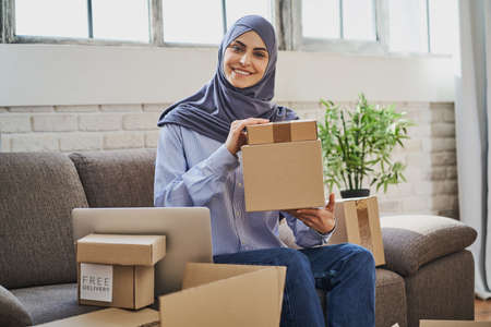 Muslim woman sitting on the couch with a laptop and showing cardboard boxesの写真素材