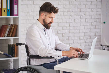Concetrated office employee sitting on a wheelchair and working on a laptopの写真素材