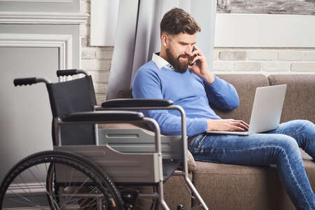 Disabled businessman sitting on a couch, working on a laptop, talking on a phoneの写真素材