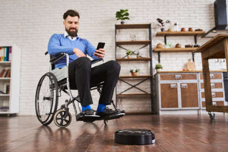 Man sitting in a wheelchair and using a robotic vacuum connected with a phoneの写真素材