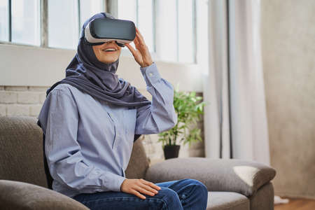 Joyful Arabian woman sitting on a couch, wearing a VR helmet and smilingの写真素材