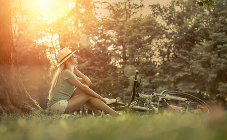 Woman sitting under sun light at day near her bicycle in the parkの写真素材