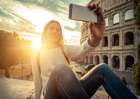 Woman tourist selfie with phone camera in hands near the Coliseum in Rome under sunlight and blue sky. Famous popular touristic place in the world.の写真素材