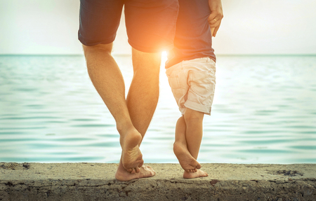 Father and son stay on the sea beach under sunlight at summer time. Close-up foots view on the sealine background at sunny day.の写真素材