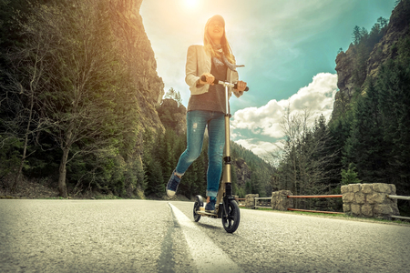 Woman on her  kick scooter on the road around mountains, under sunlight at sunny summer day.の写真素材
