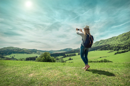 Woman walking around the mountains with her phone and shootinh beautiful green fields view under sunlight on summer day.の写真素材