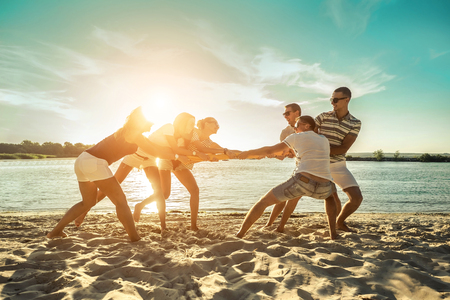 Friends funny tug of war on the beach under sunset sunlight in summer sunny day.の写真素材