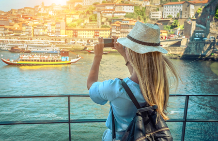 Happy blonde woman - tourist shot on her smartphone camera beautiful city view. with ships on the river in sunny day.の写真素材
