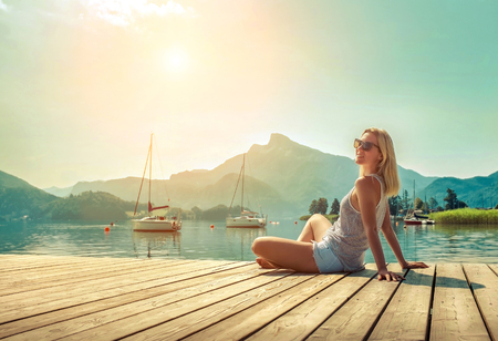 Happiness blonde woman seat on the wooden pier near the mountain lake under sunlight in morning time in summer.の写真素材