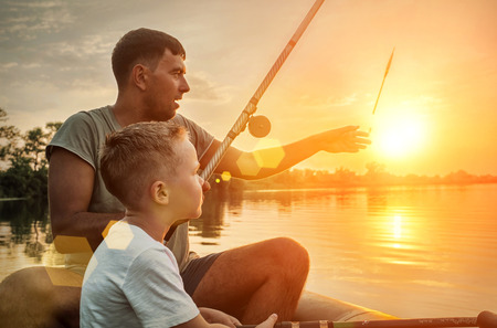 Happy Father and Son together fishing from a boat at sunset time in summer day under beautiful sky on the lake.の写真素材
