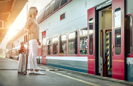 Blonde woman with her luggage stay near the red train on the peron os rail station under sun light at sunny day.