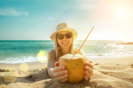 Happy Young Tourist  Smiling Caucasian Woman in hat with coconut in her hands on beach at sunny day. Beautiful Sunset light on coastline.の写真素材