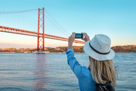 Young traveler woman with smartphone takes pictures in Lisbon of city near the bridge under sunlight in sunny summer day.の写真素材