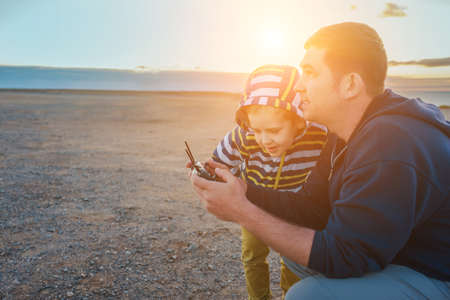 Father and son controlling fly drone on the seashore at sunset in summer day. Freedom, Family,...