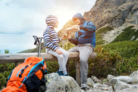 Happy hiker traveler father and son sit on the top of mountain, talking and drink hot tea a thermos....