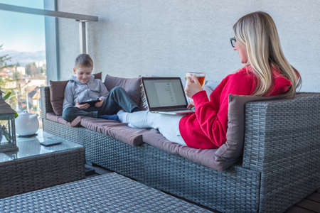 Stay at home mom. Mother and son spend free time on terrace. Woman working, child using smartphone....
