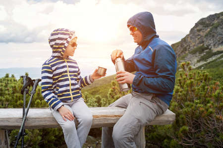Happy hiker traveler father and son sit on the top of mountain, talking and drink hot tea a thermos. Mountains landscape view. Holidays, fall, travel day.の写真素材