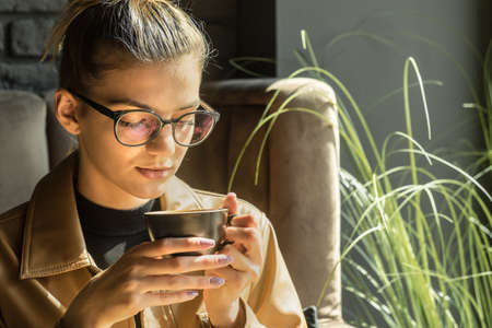 Young woman drinking coffee cup and sitting in cafe. Girl in glasses pauses after work.の写真素材