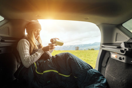 Girl resting in her car. Woman hiker, hiking backpacker traveler camper in sleeping bag, drinking...