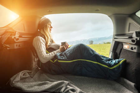 Girl resting in her car. Woman hiker, hiking backpacker traveler camper in sleeping bag, drinking hot tea and relaxing on top of mountain. Health care, authenticity, sense of balance and calmness.の写真素材