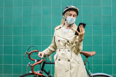 Woman in protective maskin, in helmet using her smartphone device. Female go to work on his sport retro bike. Girl standing on green background.の写真素材