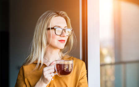 Beautiful smiling Businesswoman in glasses standing near the window and drinking tea cup. Female pretty face, work from home, social distancing, alone at home in office. close up front portraitの写真素材