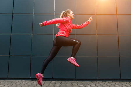 Young woman runner, jump on sidewalk on modern background. Female model in sportswear exercising...