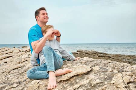 Happy family, father and son bonding, sitting on stone by the sea looking at view enjoying summer vacation. Togetherness Friendly conceptの写真素材
