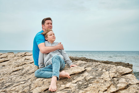 Happy family, father and son bonding, sitting on stone by the sea looking at view enjoying summer vacation. Togetherness Friendly conceptの写真素材