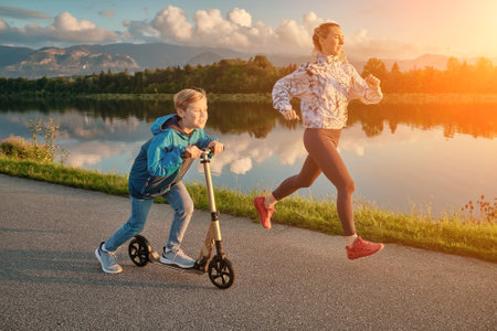 Happy mother and son go in sports outdoors. Boy rides scooter, mom runs on sunny day. Silhouette family at sunset. Fresh air. Health care, authenticity, sense of balance and calmness.の写真素材