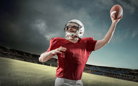 American football sportsman player with ball in action on stadium under lights of background. Sport, proud footballer in white helmet and red t-shirt ready to play.の写真素材