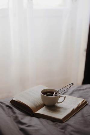 Cozy home still life: cup of hot tea with sieve and opened book on grey bed. The concept of coziness and reading. Relaxing morning. Winter holidays, free copy spaceの写真素材