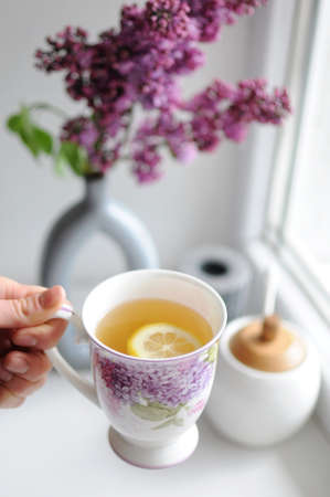 Top view concept of woman hand holding a cup of tea. Lilac flowers on backgroundの写真素材