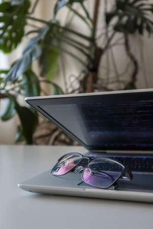 Mans glasses are lying on a laptop with code at home. Home office with plantsの写真素材