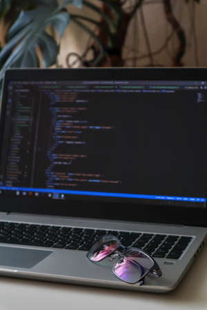 Mans glasses are lying on a laptop with code at home. Home office with plantsの写真素材