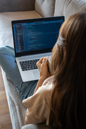 A young woman programmer is coding. A woman is drinking tea while working on a laptop and sitting on...
