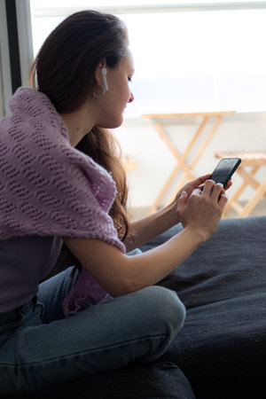 A young woman in headphones is sitting on a couch sofa at home and using her phone.