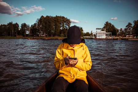 Young woman in a yellow raincoat and cap sailing on a boat and looking at her phone. A person is...
