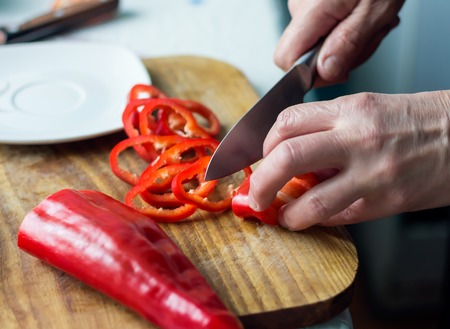 Woman is chopping red bell pepper with knife on a wooden board in the kitchen. Female hands cutting vegetables for salad. Healthy natural food with vitaminsの写真素材