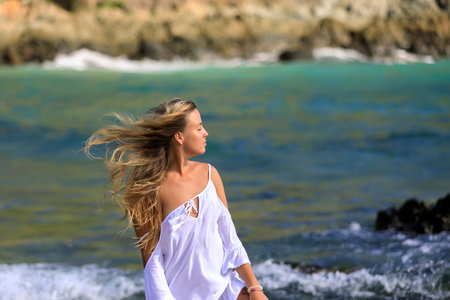 A young beautiful girl looks at the sea, the wind develops her blond hair. White dress, blue water., Beautiful view.の写真素材